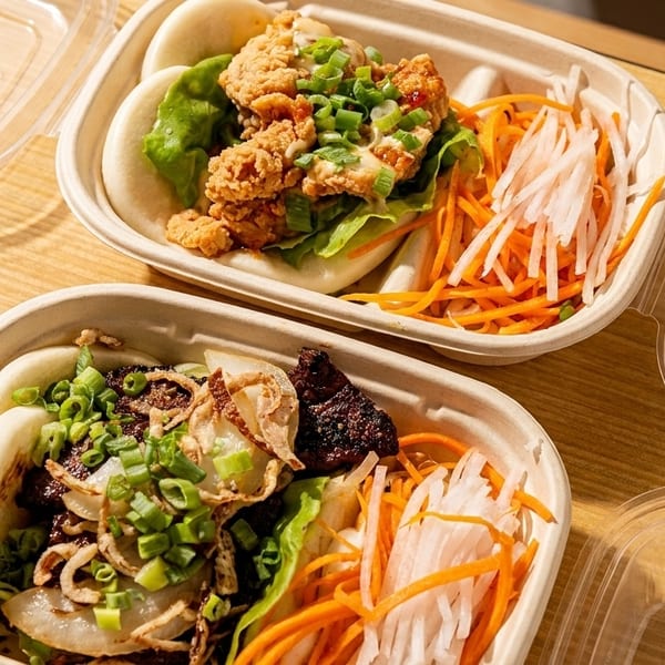 Two takeout containers with bao buns, one with fried chicken, the other with beef and onions.