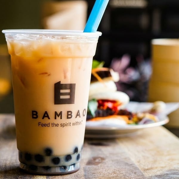 A BambaO bubble tea with a blue straw, bao buns, and a bamboo steamer on a wooden table.