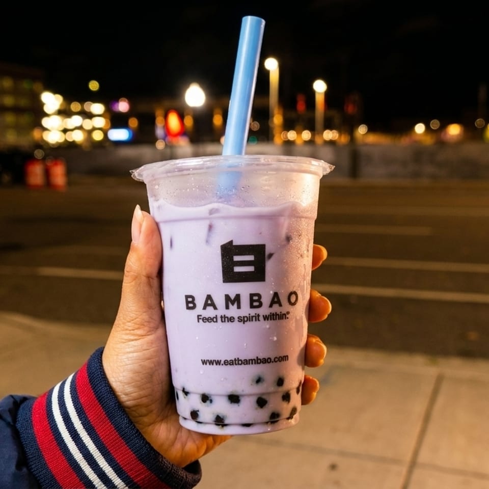 Hand holding a Bambao taro bubble tea with a blue straw against a blurred city night background.